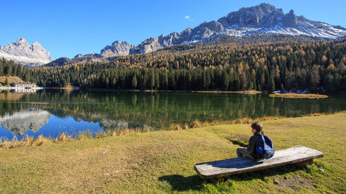 Rear view of boy sitting on bench in front of lake with mountains reflection