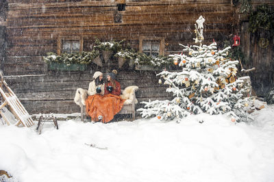 Friends sitting on bench by christmas tree in front of mountain hut