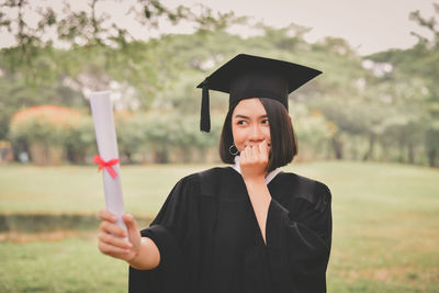 Happy young woman in graduation gown standing on field