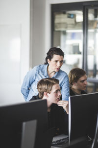 Confident female mature teacher standing by teenage student while looking at computer monitor in classroom