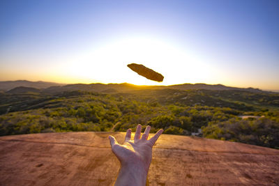 Cropped image of hand throwing stone against clear sky during sunset