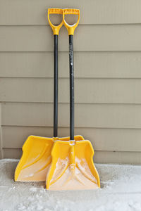 Two yellow and black snow shovels are dusted in snow and leaning against a wall ready for use. 