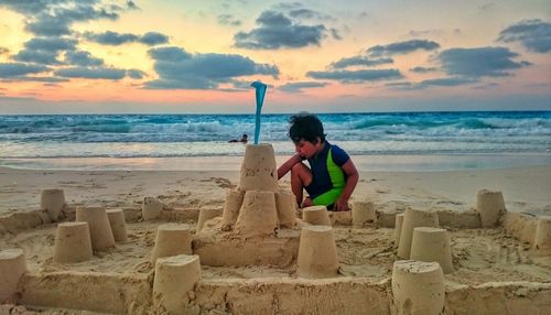 Boy making sandcastle at beach against sky during sunset