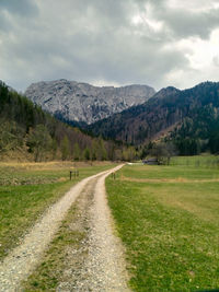 Dirt road amidst green landscape and mountains against sky
