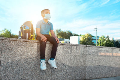 Boy, schoolboy in protective mask sits and wait near school in sunny september day.