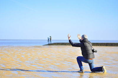 Rear view of man gesturing while kneeling at beach