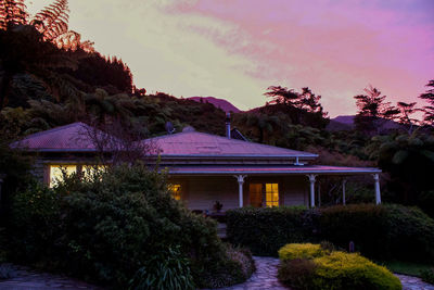 Illuminated building by trees against sky at sunset