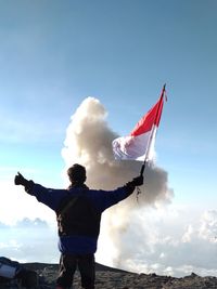 Rear view of man with arms outstretched holding indonesian flag while standing on mountain against sky