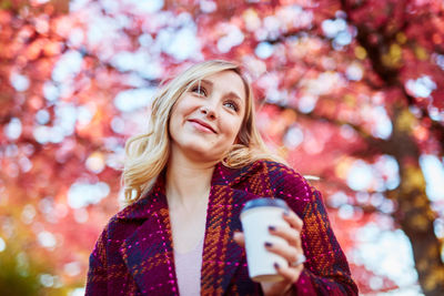 Young woman holding coffee while standing against autumn trees