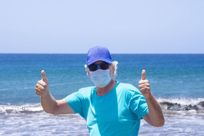 Senior man with protective mask showing thumbs up standing by sea against sky