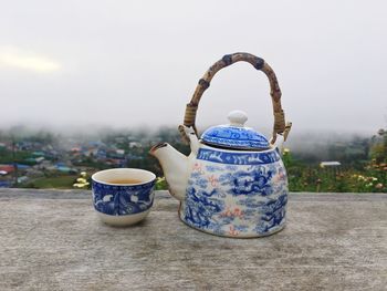 Close-up of kettle with tea cup on table against cloudy sky