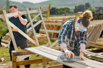 Focused female sanding down wooden planks with special electric orbit sander while working in countryside near man carrying wooden construction