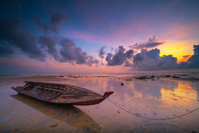 Abandoned rowboat moored on shore at beach against dramatic sky during sunset