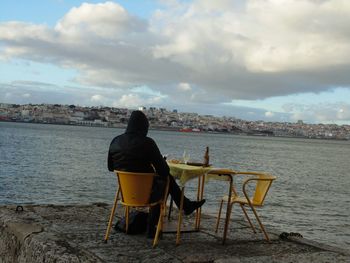 Rear view of man sitting on chair by river against sky