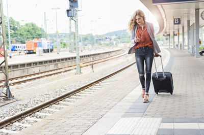 Young woman with wheeled luggage looking at her smartphone while walking at platform