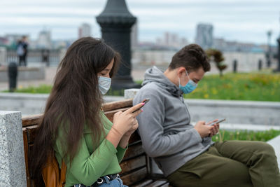  a guy and a girl are sitting on a bench at a distance from each other with masks on their faces 