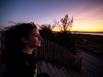 Side view of woman looking away while sitting on steps against sky during sunset