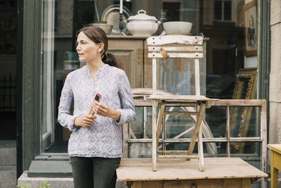 Woman looking away while standing by chair and table outside store