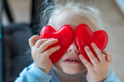 A toddler girl is holding two red hearts at the eyes