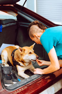 Side view of crop female in casual wear with mohawk hairstyle feeding dog sitting in car trunk at outside of house
