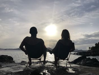 Rear view of people sitting on rock by sea against sky