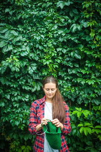 A young dark-haired woman in a plaid red shirt and blue jeans stands on a background of green wall 