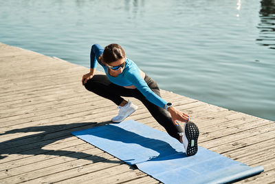 From above active slim female in sportive outfit standing on mat and doing stretching exercise for legs while training on wooden pier near river