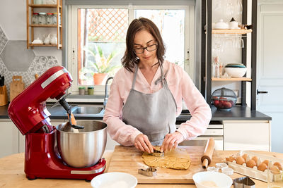 Woman wearing apron baking cookies in cozy kitchen. housewife cuts out cookies in shape of a heart