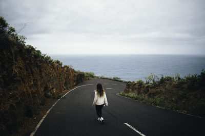 Back view of anonymous female traveler walking on asphalt road towards sea on cloudy day in la palma, spain