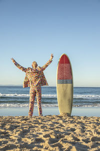 Man in colorful suit standing with arms outstretched by surfboard at beach