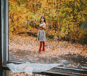 Full length portrait of woman against autumn trees on field seen through broken glass window