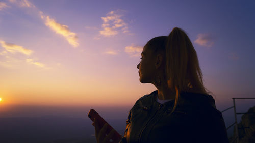 Young woman holding mobile phone while standing against sky during sunset