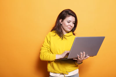 Young woman using laptop while standing against yellow background