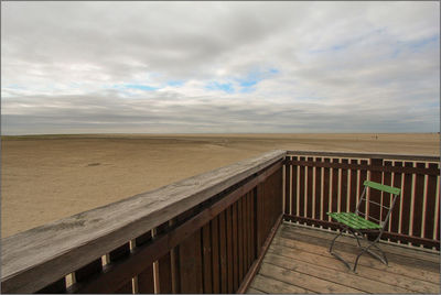 Empty chair in balcony at beach against cloudy sky