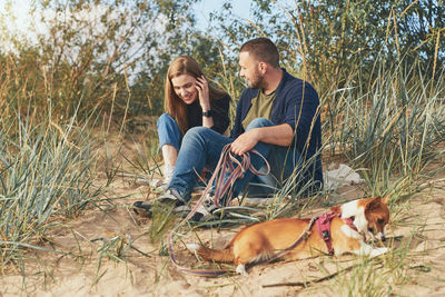 Young happy couple of man and woman with corgi dog sit at sand. two persons in casual clothes