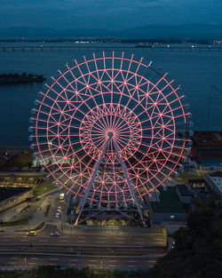 Illuminated ferris wheel by sea against sky in city