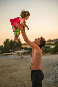 Shirtless father carrying daughter while standing at beach against sky