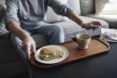 Midsection of senior man with sandwich and cup sitting on sofa at home