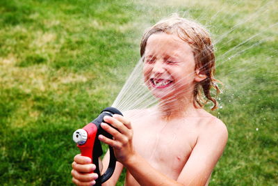 Shirtless boy splashing water from garden hose on face in back yard