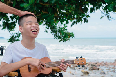 Close-up of smiling boy sitting on wheelchair and playing guitar against sea