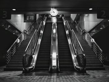 Black and white image of a clock above a  shiny escalator with staircase at each sides