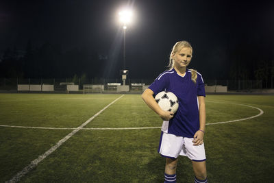 Portrait of confident girl standing with ball on soccer field against sky at night