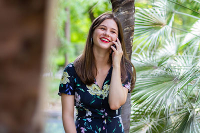 Beautiful young woman using phone while standing against tree trunk