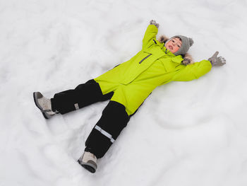 Smiling boy in green jumpsuit is making snow angel shape on snow. joyful child playing outdoors