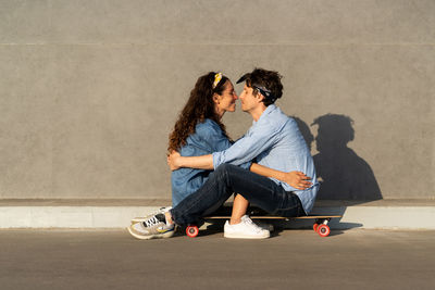 Trendy young couple sit on longboard on sunny city street happy smiling embrace enjoy time together