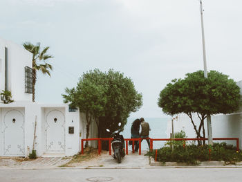 Rear view of friends sitting on railing amidst buildings against sea