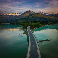 Road leading towards mountains by lake against sky during sunset