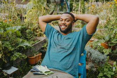 Portrait of smiling male volunteer with hands behind head sitting in community garden