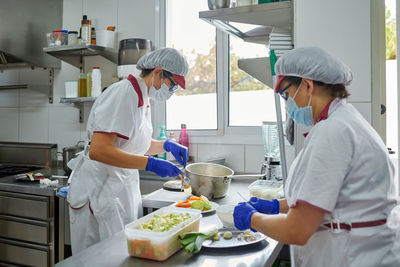 Side view of unrecognizable female cooks in protective masks and gloves preparing meal for patients while working in hospital kitchen during coronavirus pandemic