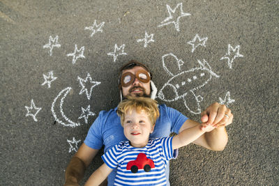 Portrait of mature man wearing pilot hat and his little son lying on asphalt painted with airplane, moon and stars